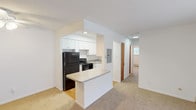 This single-floor residence offers a practical and comfortable layout designed for everyday living. The kitchen is a standout feature, showcasing crisp white cabinetry, granite-style countertops, hardwood flooring, and black appliances including a refrigerator, range, and dishwasher. A peninsula counter opens seamlessly to the adjacent living area, creating an ideal flow for daily life and entertaining. The living room is generously proportioned, filled with natural light from a large sliding glass door fitted with vertical blinds, and includes a wall-mounted air conditioning unit and baseboard heating for year-round comfort. Soft carpet extends throughout the living and bedroom areas, adding warmth underfoot. Ceiling fans with integrated lighting are found in both the living room and bedrooms, enhancing comfort and air circulation. The bedrooms are well-sized, with clean white walls, carpeted floors, and natural light from windows overlooking greenery. Warm wood-toned interior doors add a classic touch throughout the unit. The bathroom features a full soaking tub and shower combination with a distinctive blue mosaic tile surround, complemented by neutral tile flooring. A hallway connects the main living spaces to the bedrooms and bathroom, with convenient closet access along the way.