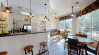 This single-floor cafe combines a warm, inviting atmosphere with practical design. The herringbone-patterned brick flooring runs throughout, lending a classic, rustic character to the entire space. A prominent service counter with a dark granite top separates the fully equipped kitchen from the dining area. The kitchen features gray cabinetry, a stainless steel refrigerator, a range with a wall-mounted range hood, and decorative pendant lighting overhead, creating a functional yet charming culinary workspace.

The dining area is furnished with a mix of round wooden tables paired with cushioned chairs and iron-accented seating, accommodating guests comfortably. Adjustable bar stools line the service counter, offering an additional casual seating option. Ceiling fans are positioned throughout, maintaining airflow across the space.

Large windows dressed with draped brown valances flood the interior with abundant natural light and frame picturesque views of a landscaped outdoor area featuring a pool, stone columns, pergola structures, and mature trees. The bright, airy ambiance created by these expansive windows gives the cafe an open, welcoming feel. Overall, the space balances a cozy, cottage-inspired aesthetic with functional amenities suited for a café setting.