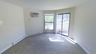 This single-floor space centers around a carpeted living area that benefits from generous natural light streaming through a large picture window and a sliding glass door. The sliding door provides direct access to an outdoor area, offering views of surrounding greenery and creating a seamless connection between indoor and outdoor living. The neutral-toned walls and light carpet contribute to a clean, understated aesthetic throughout the space. Climate control is handled by a wall-mounted air conditioning unit, complemented by baseboard heating running along the perimeter of the room — ensuring year-round comfort. The layout is open and straightforward, making the living area feel functional and adaptable to various furniture arrangements. The combination of the picture window and sliding glass door floods the space with sunlight during the day, enhancing the overall brightness. The exterior views reveal mature trees and landscaping, adding a touch of nature to the interior ambiance. Overall, the space presents a practical and comfortable living environment with key functional features already in place.