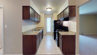 This single-floor unit offers a practical and functional layout across several rooms. The living area is generously proportioned, featuring neutral-toned carpet, light-colored walls, and large windows dressed with vertical blinds that allow ample natural light to filter in. A second carpeted room, likely a bedroom, mirrors this design with its own window overlooking greenery outdoors, flush-mount ceiling lighting, and ample wall space. The entry area connects seamlessly to the living space through a solid wood door, providing a welcoming transition into the home.

The galley-style kitchen runs along two parallel walls, featuring dark-stained wood cabinetry on both sides, light-colored countertops, and tile flooring. Black appliances, including a range and refrigerator, are integrated into the layout, and a window at the far end introduces natural light into the cooking space.

The bathroom includes a single-basin vanity with a stone-toned countertop, dark wood cabinetry below, and mirrored medicine cabinets above. Mosaic tile flooring adds texture to the space, while a built-in shelving unit visible through an adjacent doorway provides convenient storage. Overall, the unit presents a clean, no-frills interior with consistent finishes throughout.