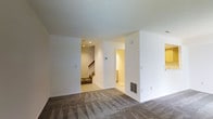 This two-story space is characterized by its clean, minimalist aesthetic with crisp white walls throughout. The main level features wall-to-wall carpeting in a neutral gray tone with a subtle pattern, lending warmth underfoot while complementing the bright, airy interior. Recessed lighting illuminates key areas, while a floor vent provides efficient climate control.

The open layout of the main level is defined by several distinct zones separated by partial walls, creating a sense of organization without fully enclosed rooms. A doorway leads to an adjacent room bathed in warm lighting, suggesting additional functional space. A staircase with wooden steps and a painted banister connects the two floors, visible through an open passageway that adds depth to the overall layout.

The ceilings are smooth and generously proportioned, contributing to the spacious feel of the interior. The neutral color palette of whites and warm beiges is carried consistently throughout, creating a cohesive and welcoming atmosphere. Overall, the space offers a practical, well-lit environment with a straightforward design suited for versatile use across both levels.