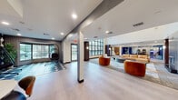 This contemporary single-floor amenity space is thoughtfully designed to foster community and comfort. The entry lobby sets an inviting tone with rich hardwood flooring, large glass doors that allow ample natural light, and a built-in mailbox center along an accent wall.

The expansive main lounge serves as the heart of the space, featuring soaring ceilings, light hardwood flooring, and a dramatic floor-to-ceiling tiled fireplace as a central focal point. Comfortable cognac leather and velvet seating arrangements are paired with a marble-patterned area rug, creating a warm yet modern aesthetic. Dark wood columns punctuate the open layout, adding architectural definition throughout.

A cozy sitting nook offers a more intimate retreat, furnished with orange velvet accent chairs set against a rich wood panel wall and floor-to-ceiling grid windows overlooking an outdoor pool area.

The game room is a standout entertainment feature, complete with polished hardwood floors, skee-ball lanes, a classic arcade cabinet, and a bar-height game table — ideal for recreation and social gatherings.

An additional lounge area near the entry features a suspended egg chair atop a geometric tile inlay, adding a playful design element. Throughout the space, recessed lighting, wall sconces, and large windows maintain a bright, modern, and welcoming atmosphere.