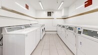 Located at 225 Van Horne Ave in Toronto's M2J neighbourhood, this shared laundry facility is a well-organized and efficiently designed amenity space. The room is finished with light-toned large-format floor tiles that lend a clean, bright aesthetic throughout. White walls are accented by a distinctive tan horizontal stripe that wraps the perimeter, adding a subtle decorative touch to the utilitarian space.

The facility is equipped with multiple top-load washing machines and front-load dryers arranged along opposing walls, providing ample capacity for residents. Each machine features a card-based payment system for convenient use. Overhead, fluorescent strip lighting ensures the space is well-illuminated at all times, complemented by a window that allows natural light to filter in.

A metal bench is positioned along one wall, offering a comfortable waiting area for residents. Additional features include a recycling bin, wall-mounted informational signage, a dispenser unit, and a baseboard heating system to maintain comfort year-round. Security cameras are installed at ceiling level, ensuring the safety of the space. A dedicated exit door provides direct access in and out of the facility. Overall, the laundry room is clean, practical, and thoughtfully appointed to serve the needs of the building's residents.