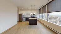 Located in the heart of Toledo, Ohio, this modern single-floor unit sits within a high-rise building on N St Clair Street, offering an impressive blend of contemporary finishes and elevated urban living. The open-concept living and kitchen area is anchored by light hardwood-style flooring and floor-to-ceiling windows fitted with sleek roller shades, providing sweeping panoramic views of the Toledo skyline and surrounding cityscape. The kitchen is well-appointed with dark espresso cabinetry contrasted by crisp white upper cabinets, granite countertops, a central island with pendant lighting, and a full suite of stainless steel appliances including a refrigerator, electric range, microwave, and dishwasher. The carpeted bedrooms offer a quiet retreat, each featuring generous closet space with sliding doors and dark-trimmed door frames that carry throughout the unit's interior design. The hallway connects the living spaces efficiently, with warm wood-tone flooring extending from the kitchen into the corridor. The bathrooms are clean and functional, each featuring a tub/shower combination, a dark-vanity with a large mirror, tile-style flooring, and brushed nickel fixtures. Throughout the unit, a neutral color palette, modern door hardware, and large-scale windows create a bright, cohesive, and polished atmosphere.