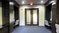 This single-floor commercial space offers a well-appointed interior designed for both functionality and a polished professional atmosphere. The central focal point is a set of dark-framed double doors with full-length glass panels, allowing visibility into the adjacent area and creating a sense of openness and flow between spaces. The doors are fitted with a door closer mechanism, reflecting practical, code-compliant design.

The flooring throughout features a blue patterned carpet in a grid motif, adding texture and a refined character to the space. Dark crown molding lines the upper perimeter of the walls, providing a strong contrast against the light-colored walls and lending a structured, finished aesthetic to the interior.

Recessed ceiling lighting ensures even, consistent illumination throughout the space, complemented by an illuminated exit sign that speaks to the space's commercial designation. On either side of the double doors, built-in desk areas with office chairs are neatly tucked into alcoves, suggesting dedicated workstation or business center functionality — ideal for guests or staff requiring computer access.

Overall, the space conveys a clean, organized, and professionally maintained environment, well-suited for a hospitality or corporate setting.