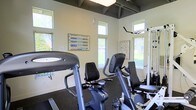 This dedicated fitness center is designed to support a comprehensive workout experience within a bright and functional single-floor layout. The space is lined with multiple windows positioned along the walls, allowing generous natural light to fill the room and providing views of the surrounding greenery outdoors — creating an open, motivating atmosphere for exercise.

The facility is equipped with a variety of cardio machines, including recumbent bikes and other cycling equipment, suitable for low-impact cardiovascular training. A cable and weight machine is positioned near the windows, offering options for strength and resistance training. Rubber flooring throughout the space provides durability and safety underfoot during workouts.

Instructional fitness charts are mounted on the walls, offering exercise guidance and workout references for users. The ceiling features exposed dark beams that add a structured, industrial character to the otherwise clean and neutral interior, with light-colored walls contributing to the bright and airy feel of the room. The overall design of the space strikes a balance between practicality and comfort, making it a well-rounded fitness facility suited for a range of exercise routines.