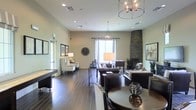 This single-floor space presents an expansive, open-plan layout designed for both relaxation and socializing. Rich, dark hardwood floors run throughout, anchoring the room's sophisticated aesthetic and providing a warm contrast to the light, neutral-toned walls. The soaring ceilings amplify the sense of openness and are adorned with two striking wrought-iron chandeliers that cast a warm, ambient glow across the entire space.

Natural light flows in through large windows dressed with sheer white curtains, brightening the central seating area, which features plush leather sofas and upholstered chairs arranged around a dramatic floor-to-ceiling stone fireplace. The fireplace serves as a commanding focal point, adding both architectural character and a cozy atmosphere to the lounge area.

Toward the front of the space, a casual sitting nook with armchairs and a table lamp provides a quieter retreat. A game table — appearing to be a shuffleboard — is positioned along one wall, adding a recreational element to the layout. A round dining or gathering table with upholstered chairs anchors the foreground, offering additional flexible seating. Framed artwork and decorative accents, including a large wall clock and potted greenery, complete the polished yet inviting ambiance of this well-appointed communal space.