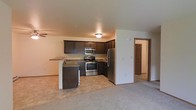 This single-floor residence offers a functional and well-appointed layout designed for comfortable everyday living. The open-concept kitchen and living area form the heart of the space, creating a seamless flow between cooking and relaxing areas. The kitchen is equipped with a full suite of stainless steel appliances, including a refrigerator, range with oven, and an over-the-range microwave. Rich, dark cabinetry provides generous storage, while the granite-style countertops add a polished touch. A breakfast bar peninsula extends into the living area, offering additional prep space and casual seating. The living area features soft carpeted flooring, creating a warm and inviting atmosphere, while the kitchen transitions to durable, easy-to-clean tile or vinyl flooring. Neutral wall tones throughout keep the space bright and versatile. Flush-mount ceiling lighting illuminates both the kitchen and adjoining areas. A hallway visible from the main living space leads to additional rooms, suggesting a well-organized and private sleeping area. Overall, the space balances practicality with modern finishes, making it an appealing and move-in-ready home.