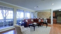 This single-floor space makes a strong first impression with its thoughtfully designed living area. The room is anchored by a bold rust-colored sectional sofa complemented by patterned accent chairs, all arranged around a dark wood coffee table atop a neutral herringbone area rug. Warm hardwood flooring runs throughout, adding richness to the overall aesthetic.

Crown molding frames the textured ceiling, lending a classic, refined character to the space. A striking stacked stone accent wall with an arched architectural detail serves as a focal point, adding texture and visual depth. Recessed lighting and a visible HVAC vent point to practical, well-considered infrastructure throughout.

Large multi-pane windows line one wall, inviting abundant natural light and offering views of an outdoor balcony or deck area with mature trees visible beyond — creating a seamless connection between the interior and the outdoors. The open layout allows the living area to flow naturally toward a dining area in the background, where white dining chairs and matching table lamps are visible.

Overall, the space combines traditional architectural elements with warm, inviting furnishings to create an atmosphere that is both comfortable and polished — ideal for everyday living and entertaining alike.
