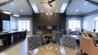 This refined single-floor space exudes sophistication through a carefully curated blend of contemporary and classic design elements. The main gathering area is anchored by a striking floor-to-ceiling stacked stone accent wall in deep charcoal tones, which serves as a dramatic focal point and adds rich texture to the room. Multiple dining tables with polished surfaces are complemented by plush, tufted wingback chairs upholstered in soft grey fabric, creating an atmosphere that is both comfortable and refined. The elevated tray ceilings add architectural depth and volume to the space, while a combination of a contemporary ceiling fan and an elegant chandelier provide layered lighting that can adapt to various moods and occasions. Recessed lighting throughout ensures the space is well-illuminated. Large windows on multiple walls flood the interior with abundant natural light, enhancing the room's open and airy feel. The warm hardwood flooring grounds the space and ties together the darker tones of the stone wall and the neutral palette of the furnishings. Overall, the space strikes a balance between grand design and inviting comfort, making it well-suited for gatherings, dining, or social events.