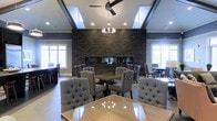 This refined single-floor space exudes sophistication through a carefully curated blend of contemporary and classic design elements. The main gathering area is anchored by a striking floor-to-ceiling stacked stone accent wall in deep charcoal tones, which serves as a dramatic focal point and adds rich texture to the room. Multiple dining tables with polished surfaces are complemented by plush, tufted wingback chairs upholstered in soft grey fabric, creating an atmosphere that is both comfortable and refined. The elevated tray ceilings add architectural depth and volume to the space, while a combination of a contemporary ceiling fan and an elegant chandelier provide layered lighting that can adapt to various moods and occasions. Recessed lighting throughout ensures the space is well-illuminated. Large windows on multiple walls flood the interior with abundant natural light, enhancing the room's open and airy feel. The warm hardwood flooring grounds the space and ties together the darker tones of the stone wall and the neutral palette of the furnishings. Overall, the space strikes a balance between grand design and inviting comfort, making it well-suited for gatherings, dining, or social events.