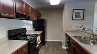 This single-floor residence offers a thoughtfully designed layout centered around a functional and stylish kitchen. The kitchen is a standout feature, showcasing rich, dark-stained wood cabinetry that lines both sides of the space, providing generous storage and a warm, classic aesthetic. Black appliances — including an electric range, refrigerator, and dishwasher — complement the deep cabinetry tones and create a cohesive, modern look. Light-toned countertops offer a subtle contrast against the darker elements, adding visual balance to the room. The flooring throughout is a durable hardwood-style material in a deep, warm brown finish, lending a grounded and inviting feel underfoot. Neutral beige walls keep the space feeling open and versatile, allowing the cabinetry and appliances to take center stage. A white paneled door leads further into the residence, hinting at additional living areas beyond the kitchen. Overhead, flush-mount lighting provides even, practical illumination across the workspace. A framed piece of wall art adds a personal touch near the entryway, contributing warmth and character. Overall, the space presents a clean, well-maintained interior with a cohesive design palette suited to comfortable everyday living.