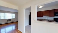 This single-floor residence at Leonardine Gardens offers a clean, functional layout with consistent design elements throughout. Warm hardwood flooring flows through the living, dining, and bedroom areas, complementing the neutral white walls and creating a bright, cohesive atmosphere.

The kitchen is outfitted with rich cherry wood cabinetry extending to the upper walls, offering generous storage capacity. Granite-style countertops, a stainless steel sink, and a black refrigerator complete the space, which features light tile flooring. A pass-through opening connects the kitchen to the adjacent hallway, enhancing the sense of flow between spaces.

The dining area is a standout feature, highlighted by a classic chandelier and a sliding glass door that floods the room with natural light and provides direct access to an outdoor area with green views. Baseboard heating runs along the walls throughout the unit, ensuring year-round comfort.

The bedrooms are well-proportioned, each featuring polished hardwood floors, wall-unit air conditioning, and individual closets with hanging rods. Multiple rooms benefit from natural light through standard windows with outdoor views.

The bathroom includes a full tub-shower combination finished with clean white wall tile. Overall, the residence presents a straightforward, well-maintained interior suitable for comfortable everyday living.