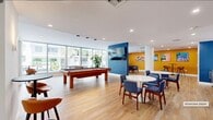 Located at 120 N Compass Way in Dania Beach, Florida, this generously sized amenity lounge offers a dynamic and well-appointed gathering space designed for both recreation and relaxation. The open-plan layout is unified by warm wood-look flooring and crisp white ceilings with recessed lighting, creating a bright and welcoming atmosphere throughout.

A central billiards table anchors the main recreation area, illuminated by a trio of modern pendant lights and flanked by floor-to-ceiling windows that invite abundant natural light and views of the surrounding landscape. A bold blue accent column mounted with a large flat-screen TV serves as a visual focal point, dividing the space into distinct activity zones while maintaining an open feel.

The seating area features round white tables paired with upholstered blue chairs, ideal for casual dining or socializing. A counter-height bar ledge set against a textured blue grasscloth accent wall provides additional seating with caramel-toned bar stools. The adjacent kitchenette is equipped with cabinetry, a refrigerator, and a built-in microwave, supporting everyday use.

Vibrant mustard-yellow walls adorned with tropical palm tree artwork add a lively, Florida-inspired energy to the lounge. A glass-walled private room offers a quieter, enclosed option within the space. Large windows throughout the lounge overlook neighboring amenities, reinforcing the connected, community-oriented character of this well-designed facility.
