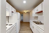 This single-floor residential unit offers a well-organized layout across several distinct spaces. The galley-style kitchen is a functional highlight, featuring white cabinetry with brushed hardware, light stone-pattern countertops, a stainless steel sink, and white appliances including a refrigerator and electric range. Recessed lighting illuminates the space, which transitions via a hallway to the rest of the unit. The living areas are finished with wood-look plank flooring and neutral-toned walls, creating a clean, cohesive aesthetic. A ceiling fan serves the main living space, and a wall-mounted air conditioning unit provides supplemental climate control. Baseboard heating runs along the walls throughout, complementing the wall unit. The bedrooms feature plush carpeting, ceiling fans, and windows dressed with horizontal blinds, allowing natural light while maintaining privacy. One bedroom includes two closets and a wall AC unit. The full bathroom presents a tub and shower combination with a white surround, wood-look flooring, and a white vanity with understated hardware. Throughout the unit, a consistent neutral color palette and clean finishes contribute to a bright, move-in-ready atmosphere.