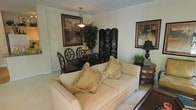 This single-floor space presents an open layout that seamlessly integrates a living area and dining space within one cohesive environment. The living area is anchored by a cream-toned sofa adorned with fringed decorative pillows in warm gold and earth tones, lending a classic, elegant feel. A dark wood side table with curved drawers sits nearby, topped with a table lamp and small greenery, adding both function and warmth to the corner.

The dining area, positioned toward the rear of the space, features a dark wood table surrounded by ornate, high-backed chairs with lattice-style detailing. A pendant light hangs overhead, illuminating the table and a decorative centerpiece. A large framed artwork on the wall and a lush potted plant contribute to the richness of the setting, while a decorative folding screen with intricate ironwork detailing serves as a stylish room divider.

The walls are finished in a neutral tone, complemented by light carpeting throughout, creating a bright and cohesive backdrop. An additional framed piece on the right wall adds a finishing artistic touch. Overall, the space conveys a traditionally styled, well-appointed interior with layered décor and a welcoming atmosphere.