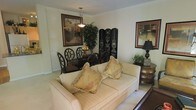 This single-floor space presents an open layout that seamlessly integrates a living area and dining space within one cohesive environment. The living area is anchored by a cream-toned sofa adorned with fringed decorative pillows in warm gold and earth tones, lending a classic, elegant feel. A dark wood side table with curved drawers sits nearby, topped with a table lamp and small greenery, adding both function and warmth to the corner.

The dining area, positioned toward the rear of the space, features a dark wood table surrounded by ornate, high-backed chairs with lattice-style detailing. A pendant light hangs overhead, illuminating the table and a decorative centerpiece. A large framed artwork on the wall and a lush potted plant contribute to the richness of the setting, while a decorative folding screen with intricate ironwork detailing serves as a stylish room divider.

The walls are finished in a neutral tone, complemented by light carpeting throughout, creating a bright and cohesive backdrop. An additional framed piece on the right wall adds a finishing artistic touch. Overall, the space conveys a traditionally styled, well-appointed interior with layered décor and a welcoming atmosphere.
