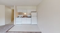 This single-floor unit offers a straightforward, functional layout with a clean, neutral aesthetic throughout. The living area is generously sized, featuring soft wall-to-wall carpeting, a textured ceiling, and a large window that allows natural light to fill the room. A baseboard heating unit and wall-mounted air conditioner provide year-round climate control, while the outdoor view adds a pleasant sense of openness.

The kitchen is efficiently designed with white cabinetry extending to the ceiling for ample storage, white appliances including a range, microwave, and full-size refrigerator, and a mosaic tile backsplash that adds a subtle decorative touch. The tile flooring transitions seamlessly from the kitchen into the adjoining areas.

The bedroom is comfortably proportioned with the same plush carpeting found in the living area, neutral walls, and a reach-in closet with a full-width rod for organized storage. A textured ceiling is consistent throughout the space.

The bathroom features a white vanity with a marble-patterned countertop, a tri-light fixture above a framed mirror, and a tiled tub-shower combination. The all-white palette creates a clean, bright atmosphere. Overall, the unit presents a cohesive, move-in-ready space with practical finishes.