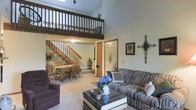 This two-story home offers a thoughtfully designed layout that balances open living spaces with functional flow. The main level centers around a spacious living room highlighted by dramatic vaulted ceilings that draw the eye upward, amplifying the sense of openness and airiness throughout. A wood-railed loft on the upper level overlooks the living area below, adding architectural depth and a sense of connectivity between floors. Natural light filters in through a skylight above, brightening the interior and enhancing the welcoming atmosphere.

The living room is comfortably appointed and opens seamlessly to an adjacent dining area, which features a classic wooden dining set suitable for family gatherings. A wood staircase with matching balustrades leads to the upper level, serving as both a functional and decorative element of the home. Neutral-toned carpeting runs throughout the main living areas, contributing to a warm and cohesive aesthetic. Wall decor and accents add a personal, homey character to the space.

A front entry door is visible from the main living area, suggesting a practical and open layout near the entryway. The overall ambiance of the home is cozy and inviting, with classic design elements, warm wood tones, and generous vertical space coming together to create a comfortable, livable environment.
