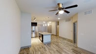 This single-story residence in Frisco, Texas, showcases a thoughtfully designed open-concept layout that seamlessly connects the kitchen and living areas. The kitchen serves as the focal point of the space, centered around a white-finished island topped with light granite or stone countertops and equipped with a built-in sink. Stainless steel appliances, including a French door refrigerator and dishwasher, lend a contemporary and functional feel to the cooking space. Dark cabinetry along the back wall provides a striking contrast to the lighter tones throughout. Pendant lighting hangs above the island, complementing the recessed lighting that illuminates the space evenly. The living area features a ceiling fan with integrated lighting, contributing to both comfort and style. Wood-look tile flooring flows consistently throughout, adding warmth while remaining easy to maintain. Neutral wall tones create a clean, versatile backdrop suitable for a variety of interior styles. Multiple doorways branch off from the main living space, suggesting well-organized access to additional rooms. The overall ambiance is bright, modern, and move-in ready.