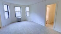 This single-floor space offers a clean and functional layout with a welcoming, light-filled main living area. The room is finished with neutral wall-to-wall carpeting that adds warmth underfoot, while crisp white walls create a bright, airy ambiance throughout. A pair of double-hung windows arranged in a bay-style configuration allows natural light to pour in from multiple angles, framing views of the surrounding neighborhood and lending the room an open, spacious feel. A classic cast-iron radiator positioned beneath the windows provides traditional radiant heating, a hallmark of well-built older construction. Clean white baseboards and trim detail the perimeter of the room, adding a subtle architectural finish consistent with the building's character. An open doorway leads directly into an adjoining bathroom, enhancing the flow and accessibility of the space. The bathroom, visible from the main room, features light-toned finishes that complement the overall neutral palette of the unit. The straightforward, uncluttered layout of the space makes it highly adaptable, offering flexibility for a variety of furnishing arrangements. Overall, the space combines classic architectural details with practical functionality.