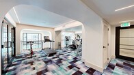 Located in Scottsdale, Arizona, this fitness center offers a thoughtfully designed two-room workout facility that balances both cardio and functional training needs. The space features a clean, contemporary aesthetic defined by elegant arched doorways, crisp white walls, and linear LED accent lighting that traces the ceiling perimeters throughout. The main exercise room is finished with a bold, multi-toned geometric carpet in shades of teal, navy, and brown, providing a vibrant visual anchor beneath an array of cardio equipment including treadmills, ellipticals, stair climbers, and stationary bikes. Large windows and glass-paneled doors open to views of an outdoor pool area, framed by decorative wrought iron railings that lend an upscale residential character to the space. A built-in dark wood cabinet with decorative accents adds a refined touch to one corner. The second exercise room transitions to warm hardwood flooring and is dedicated to cycling, housing multiple stationary bikes with individual screen monitors and wall-mounted fans for ventilation. Mirrored walls expand the visual depth of the space, while functional training accessories including medicine balls and resistance equipment round out the facility's offerings. Together, the two rooms create a comprehensive and polished fitness environment.