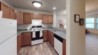 This single-floor unit offers a practical and cohesive layout across its one bedroom and one bathroom. The living room features carpet flooring, a textured ceiling, and two windows that allow natural light to filter in, creating a comfortable and inviting atmosphere. A baseboard heating unit runs along the exterior wall, providing efficient climate control.

The kitchen is efficiently designed with warm-toned wood cabinetry extending to the upper walls, offering generous storage space. Dark countertops provide a contrast to the white appliances, which include an electric range and dishwasher. Wood-look flooring runs throughout the kitchen, adding a clean, durable surface underfoot. The layout allows for easy movement and workflow between prep and cooking areas.

The bedroom is carpeted and includes a double closet with classic panel doors, providing ample storage. The room comfortably accommodates a full-sized bed and nightstand, with neutral wall tones contributing to a calm, restful environment.

The bathroom features wood-look flooring consistent with the kitchen, a wood-framed vanity with a white countertop, a wall-mounted storage cabinet above the toilet, and a shower with a curtain enclosure. Chrome fixtures and a lighted mirror complete the functional space.