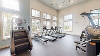 This expansive fitness center offers a generously sized, single-floor layout designed to accommodate a full range of workout routines. The space is characterized by soaring ceilings that create an open, airy atmosphere, complemented by an abundance of large, multi-pane windows along the walls that flood the interior with natural light and provide views of the surrounding outdoor environment.

The facility is divided into distinct functional zones. A dedicated cardio area houses multiple treadmills, elliptical trainers, rowing machines, and a stair climber, all positioned to take advantage of the bright, windowed walls. An adjacent cycling zone features Echelon smart bikes with digital displays for interactive workouts.

The strength training section is anchored by a comprehensive multi-station cable machine, a bench press setup, and an extensive free weights area with a full rack of dumbbells and a large mirror wall that enhances the sense of depth. Functional fitness is also well represented, with medicine balls, stability balls, an adjustable workout bench, and even a heavy tractor tire for conditioning drills.

The interior finishes include durable rubber sport flooring, recessed lighting, and ceiling fans for air circulation. Motivational fitness artwork is displayed near the upper wall. Glass-paneled double doors provide access to the outdoor area, reinforcing the connection between indoor and outdoor spaces throughout this well-appointed facility.