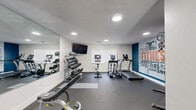 This fitness center is a thoughtfully designed, single-room workout space that maximizes its compact footprint to deliver a comprehensive exercise experience. The room features durable dark rubber flooring throughout, white walls, and a textured ceiling fitted with recessed lighting that keeps the space bright and energizing. A large wall-mounted mirror along one side visually expands the room while allowing users to monitor their form during workouts. The centerpiece of the space is a bold, colorful fitness-themed typographic mural adorning the far accent wall in blue, white, and orange — adding a dynamic, motivational atmosphere. Equipment is well-organized and varied, including treadmills, an elliptical trainer, a stationary spin bike, a multi-function cable weight machine, an adjustable weight bench, and a full rack of dumbbells and kettlebells in graduated sizes. A wall-mounted flat-screen television provides entertainment during cardio sessions. Natural light enters through a window fitted with horizontal blinds, complementing the overhead lighting. A storage rack with exercise accessories, including a stability ball and yoga mats, rounds out the amenities. The space is accessible via a dedicated entry door with emergency exit signage, making it suitable for a shared community or amenity facility.