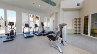 This single-floor fitness center is designed to deliver a comprehensive workout experience within a bright, open, and thoughtfully appointed space. The facility boasts soaring vaulted ceilings accented by decorative teal chevron detailing, creating an airy and energizing atmosphere throughout.

The main workout area is generously sized and outfitted with a diverse range of equipment, including multiple treadmills, an elliptical trainer, a stationary cycle, a leg press machine, a cable resistance machine, a squat rack, and a full dumbbell rack. Floor-to-ceiling mirrors line one wall, amplifying the sense of space and allowing users to monitor their form, while wall-mounted flat-screen TVs provide entertainment during workouts.

Large windows along multiple walls flood the interior with natural light and offer views of the surrounding outdoor area, which appears to include a pool. Neutral-toned carpet flooring spans the exercise space, offering comfort underfoot. Built-in sage green cabinetry and open shelving add a refined, residential touch to the facility.

A separate lounge or activity room features patterned tile flooring, seating, a small table, a pegboard wall, and a mounted television, providing a versatile space for relaxation or group activities. A ceiling fan enhances ventilation in the strength training zone, rounding out this well-appointed community fitness facility.