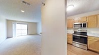 This single-floor space is thoughtfully laid out with distinct living and kitchen areas separated by a dividing wall. The living area features wall-to-wall carpeting in a neutral tone, creating a comfortable and cohesive atmosphere. A large window at the far end of the room allows natural light to filter in and provides a view of an outdoor balcony or patio, enhancing the sense of openness in the space. A textured ceiling adds a subtle visual element throughout.

The kitchen, positioned adjacent to the living area, features warm-toned oak cabinetry that provides ample storage space. Stainless steel appliances, including an electric range and an over-the-range microwave, give the kitchen a modern and functional character. Light-toned countertops complement the cabinetry, while durable hard surface flooring in the kitchen contrasts with the carpeting in the living space, clearly delineating the two areas. Overhead lighting in both areas ensures the space remains well-lit throughout the day. Overall, the layout offers a practical and comfortable living environment.