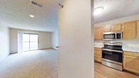 This single-floor space is thoughtfully laid out with distinct living and kitchen areas separated by a dividing wall. The living area features wall-to-wall carpeting in a neutral tone, creating a comfortable and cohesive atmosphere. A large window at the far end of the room allows natural light to filter in and provides a view of an outdoor balcony or patio, enhancing the sense of openness in the space. A textured ceiling adds a subtle visual element throughout.

The kitchen, positioned adjacent to the living area, features warm-toned oak cabinetry that provides ample storage space. Stainless steel appliances, including an electric range and an over-the-range microwave, give the kitchen a modern and functional character. Light-toned countertops complement the cabinetry, while durable hard surface flooring in the kitchen contrasts with the carpeting in the living space, clearly delineating the two areas. Overhead lighting in both areas ensures the space remains well-lit throughout the day. Overall, the layout offers a practical and comfortable living environment.