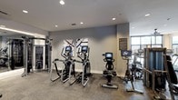 This well-appointed fitness center offers a comprehensive workout environment designed with both functionality and modern aesthetics in mind. The space is finished with neutral gray walls and recessed lighting throughout, creating a bright and energizing atmosphere conducive to exercise. Large windows allow natural light to fill portions of the room, adding to the open, welcoming feel.

The cardio area is prominently equipped with multiple elliptical trainers and stationary cycling bikes, each outfitted with individual digital display screens for on-demand workout programming and performance tracking. A rowing machine is also available, rounding out the cardiovascular options. On the opposite side of the space, a cable and pulley strength training system provides versatile resistance training capabilities.

Durable, low-pile carpet flooring covers the workout area, offering both comfort underfoot and stability during exercise. The walls are accented with decorative abstract artwork, adding a polished, curated touch to the interior. An informational signage panel is mounted near the entrance, orienting users to gym amenities and guidelines. Overall, the space strikes a balance between professional-grade equipment and an inviting, contemporary design.