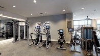 This well-appointed fitness center offers a comprehensive workout environment designed with both functionality and modern aesthetics in mind. The space is finished with neutral gray walls and recessed lighting throughout, creating a bright and energizing atmosphere conducive to exercise. Large windows allow natural light to fill portions of the room, adding to the open, welcoming feel.

The cardio area is prominently equipped with multiple elliptical trainers and stationary cycling bikes, each outfitted with individual digital display screens for on-demand workout programming and performance tracking. A rowing machine is also available, rounding out the cardiovascular options. On the opposite side of the space, a cable and pulley strength training system provides versatile resistance training capabilities.

Durable, low-pile carpet flooring covers the workout area, offering both comfort underfoot and stability during exercise. The walls are accented with decorative abstract artwork, adding a polished, curated touch to the interior. An informational signage panel is mounted near the entrance, orienting users to gym amenities and guidelines. Overall, the space strikes a balance between professional-grade equipment and an inviting, contemporary design.