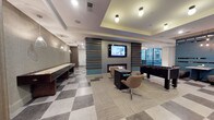 Located at 99 WestEdge Street in Charleston, South Carolina, this single-floor amenity lounge is a thoughtfully designed community space that blends entertainment and relaxation seamlessly. The entry greets visitors with a bold checkered tile floor and deep navy accent walls adorned with curated artwork, setting a sophisticated tone from the start. The expansive open layout flows effortlessly between distinct activity zones, each with its own character while maintaining a cohesive modern aesthetic. A game area anchors one side of the space, featuring a full-size billiards table, a shuffleboard table, and a foosball table, all positioned beneath stylish pendant and recessed lighting. A wall-mounted flat-screen TV and a linear fireplace framed by a striking multi-toned horizontal slat feature wall serve as a central focal point, with contemporary lounge chairs arranged nearby for comfortable viewing. Booth-style dining seating lined against a teal shiplap partition wall with pendant lighting above provides a casual gathering spot. Multiple soft seating arrangements, including large sectional sofas, accent chairs, and ottomans in neutral and teal tones, offer ample space for socializing or unwinding. Floor-to-ceiling windows along one side of the room flood the interior with natural light and provide views of the surrounding Charleston streetscape, enhancing the overall sense of openness and connectivity to the vibrant neighborhood outside.