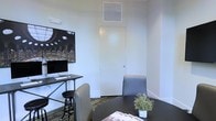 This single-floor office space offers a clean, minimalist environment well-suited for professional work and intimate meetings. The interior is defined by crisp white walls that enhance the sense of brightness and openness throughout the space. A dedicated workstation area features a sleek standing-height desk equipped with an iMac desktop computer and a black metal stool, providing an ergonomic setup for individual productivity. Adjacent to the workstation, a small meeting or consultation area includes a round dark table accompanied by upholstered gray chairs, creating an inviting setting for one-on-one conversations or small group discussions. A flush-mounted door blends seamlessly into the white wall, contributing to the space's streamlined appearance. Overhead, a ceiling-mounted HVAC vent ensures year-round climate comfort. Striped carpeting in muted tones adds a subtle texture to the floor, grounding the space with a professional feel. A small decorative plant on the table introduces a touch of greenery, softening the otherwise structured environment. Overall, the space balances functionality and understated style, making it an efficient and comfortable setting for office use.