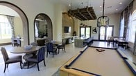 This well-appointed single-floor social and entertainment space combines thoughtful design with a variety of recreational amenities, creating an inviting environment for leisure and community gatherings. At the forefront is a full-size pool table anchored beneath a striking rustic wood ceiling fixture adorned with pendant lights, setting a warm and sophisticated tone. A textured stone accent wall serves as a dramatic backdrop for a wall-mounted flat-screen television, flanked by a media console with integrated cabinetry and beverage storage. Adjacent to the media area, an expansive island bar with a light stone countertop and high-back bar seating accommodates numerous guests, making it ideal for socializing. Natural light filters in through large windows and glass doors, brightening the polished concrete floors throughout. A curved archway with dark wood trim leads to an adjoining lounge room furnished with comfortable seating and a chandelier, offering a quieter retreat. Casual dining tables with upholstered chairs provide additional seating options. The layered lighting scheme, combining recessed fixtures, pendant clusters, and statement chandeliers, enhances the space's warm, upscale ambiance throughout.