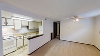 This single-story space features an open and functional layout that connects the kitchen and living area seamlessly. The kitchen is positioned to the left and is defined by a half-wall pass-through with a dark laminate countertop, creating a natural divide while maintaining an open feel. White cabinetry lines the kitchen walls, offering ample storage, and is paired with a white refrigerator and dishwasher. A fluorescent overhead light fixture illuminates the workspace, which includes a sink integrated into the countertop. The flooring in the kitchen area features light-toned tile, transitioning into wall-to-wall carpeting in the living space. The living area is generously proportioned, with white walls, white crown molding, and dark baseboards that frame the room with subtle contrast. A ceiling fan is centrally mounted, providing both light and airflow. A hallway visible in the background leads to additional rooms, suggesting further private living spaces beyond the main open area. The overall ambiance is clean, neutral, and practical, with a straightforward design that maximizes usable space throughout.