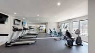 This single-floor fitness center offers a clean, modern environment designed to support a well-rounded cardio workout. The space is finished with durable dark rubber flooring throughout, providing both safety and comfort underfoot. Recessed lighting is complemented by natural light streaming in through multiple windows, giving the room a bright and energizing atmosphere.

The main workout area is generously proportioned, housing a row of Spirit-brand treadmills along one side, alongside an elliptical trainer, a recumbent bike, and an upright stationary bike. A large floor-to-ceiling mirror spans one wall, visually expanding the space and allowing users to monitor their form during exercise. Wall-mounted flat-screen televisions are positioned at multiple points around the room, keeping members entertained during workouts.

A pass-through window or interior glass panel connects the fitness room to an adjacent office or leasing area, adding a sense of openness and easy access. The walls are painted in a crisp, neutral gray-white tone that contributes to the space's clean and contemporary aesthetic. A fire extinguisher and climate control vents are also present, reflecting attention to safety and comfort. Overall, the fitness center presents a functional, well-maintained space suited for daily cardio and light exercise routines.
