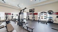 Located at 2250 Hard Scabble Road in Columbia, South Carolina, this fitness center offers a thoughtfully designed exercise space optimized for a comprehensive workout experience. The room features a neutral color palette accented by a bold red and black horizontal stripe that runs along the perimeter walls, lending the space a dynamic, energetic atmosphere. Durable dark tile flooring provides a sturdy foundation throughout, while recessed lighting and ceiling fans with integrated light fixtures ensure the room stays well-lit and well-ventilated.

Natural light filters in through multiple windows fitted with horizontal blinds, offering views of the surrounding exterior. The space is equipped with a strong lineup of cardio machines, including multiple treadmills, ellipticals, and a stationary spin bike with an integrated display screen. A stair climber rounds out the cardio offerings. On the strength training side, the room features a multi-station cable machine, a squat rack with weight plates, a dedicated dumbbell rack stocked with a full range of free weights, medicine balls, and a weight bench. Multiple wall-mounted flat-screen televisions are positioned throughout for entertainment during workouts. A sanitizing station is also present, underscoring the facility's attention to cleanliness and user comfort.