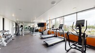 Located at 100 Citronica Ln in Lemon Grove, California, this single-level fitness center is designed to support a comprehensive workout experience. The space features a modern, open layout with a clean aesthetic defined by white walls accented by bold orange panels, rubber-speckled flooring for durability and safety, and recessed lighting throughout.

Along one wall, expansive floor-to-ceiling windows flood the cardio zone with natural light and provide views of the surrounding streetscape and palm trees, creating an energizing environment. This area is outfitted with multiple treadmills, elliptical trainers, stair climbers, and stationary bikes, each equipped with individual digital screens.

The strength training section is equally well-appointed, featuring a Hoist cable machine, a leg press, kettlebells in varying weights, medicine balls, and foam rollers neatly organized on tiered racks. Large wall-mounted mirrors extend the visual depth of the space and allow users to monitor their form. Flat-screen televisions are mounted throughout, adding entertainment options during workouts.

The facility is accessible via a glass-paneled door and offers a well-organized, distraction-free environment suitable for a wide range of fitness routines.
