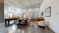 This well-appointed clubhouse spans a generous single floor, thoughtfully designed to serve as a welcoming communal gathering space. The entry foyer sets an elegant tone with large-format tile flooring, decorative medallion inlay, and classic double doors with leaded glass accents that flood the entry with natural light.

The main lounge area is anchored by soaring ceilings and rich, dark hardwood floors that extend throughout the space. Multiple seating arrangements — including plush sectional sofas, leather accent chairs, and a bold retro-patterned area rug — create inviting zones for relaxation and socializing. A wall-mounted flat-screen television and a built-in bookshelf add functional character, while recessed lighting and statement pendant fixtures provide warm, layered illumination.

A dedicated billiards area with a full pool table offers additional recreation options, complemented by a large L-shaped bench seating alcove lined with windows that bring in ample natural light.

The kitchenette features a granite-topped center island, stainless steel appliances including a French-door refrigerator, stone tile accent walls, a linear fireplace, and industrial-style pendant lighting — ideal for hosting and casual gatherings.

A separate meeting or conference area with a white table and modern wood-accented chairs rounds out this versatile and stylishly curated community space.