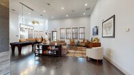This well-appointed clubhouse spans a generous single floor, thoughtfully designed to serve as a welcoming communal gathering space. The entry foyer sets an elegant tone with large-format tile flooring, decorative medallion inlay, and classic double doors with leaded glass accents that flood the entry with natural light.

The main lounge area is anchored by soaring ceilings and rich, dark hardwood floors that extend throughout the space. Multiple seating arrangements — including plush sectional sofas, leather accent chairs, and a bold retro-patterned area rug — create inviting zones for relaxation and socializing. A wall-mounted flat-screen television and a built-in bookshelf add functional character, while recessed lighting and statement pendant fixtures provide warm, layered illumination.

A dedicated billiards area with a full pool table offers additional recreation options, complemented by a large L-shaped bench seating alcove lined with windows that bring in ample natural light.

The kitchenette features a granite-topped center island, stainless steel appliances including a French-door refrigerator, stone tile accent walls, a linear fireplace, and industrial-style pendant lighting — ideal for hosting and casual gatherings.

A separate meeting or conference area with a white table and modern wood-accented chairs rounds out this versatile and stylishly curated community space.