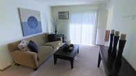 This single-floor residence centers around a well-appointed living area that serves as the heart of the space. The room is anchored by large sliding glass doors dressed with vertical blinds, allowing generous natural light to fill the space and creating a bright, airy atmosphere. Plush wall-to-wall carpeting adds warmth underfoot and contributes to the room's comfortable feel. A wall-mounted air conditioning unit ensures climate control year-round. The living area comfortably accommodates a full-sized sofa with accent pillows, a dark-finished coffee table, and a side table, demonstrating a practical and functional layout. Neutral wall tones provide a versatile backdrop, complemented by decorative wall art that adds a touch of character to the space. The overall ambiance is clean, understated, and move-in ready, with a layout that balances open sightlines with defined living zones. The combination of natural light, soft flooring, and thoughtful design elements makes this a welcoming and livable space suited to a variety of furnishing styles.