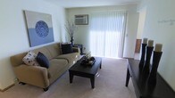 This single-floor residence centers around a well-appointed living area that serves as the heart of the space. The room is anchored by large sliding glass doors dressed with vertical blinds, allowing generous natural light to fill the space and creating a bright, airy atmosphere. Plush wall-to-wall carpeting adds warmth underfoot and contributes to the room's comfortable feel. A wall-mounted air conditioning unit ensures climate control year-round. The living area comfortably accommodates a full-sized sofa with accent pillows, a dark-finished coffee table, and a side table, demonstrating a practical and functional layout. Neutral wall tones provide a versatile backdrop, complemented by decorative wall art that adds a touch of character to the space. The overall ambiance is clean, understated, and move-in ready, with a layout that balances open sightlines with defined living zones. The combination of natural light, soft flooring, and thoughtful design elements makes this a welcoming and livable space suited to a variety of furnishing styles.