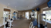 This fitness center is a single-floor facility designed to accommodate a range of workout routines. The room features warm beige walls, recessed lighting in a smooth white ceiling, and carpeted flooring that provides a comfortable surface underfoot. Natural light enters through a set of large windows and a glass-paneled door on one side, offering views of the exterior and brightening the space considerably.

The room is outfitted with a solid selection of strength training equipment, including a multi-station cable machine, weight racks, and a variety of free weights. Stability balls are neatly stored on a dedicated rack along one wall, alongside additional accessories and benches. A dumbbell rack is also visible, ensuring weights are organized and accessible. Two wall-mounted flat-screen TVs are positioned for easy viewing during workouts, enhancing the experience for users.

The layout is open and functional, allowing users to move freely between equipment stations. Recessed ceiling vents ensure proper air circulation throughout the space. Overall, the fitness center presents a clean, practical environment suitable for both light and intensive training sessions.