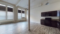 This loft-style unit showcases a compelling blend of historic architectural character and contemporary updates. The open-plan layout is anchored by striking arched windows that flood the living area with natural light, while the impressively tall ceilings — further emphasized by exposed ceiling beams, industrial-style piping, and multiple ceiling fans — create a sense of generous, airy volume throughout.

Light-toned hardwood-style flooring runs continuously across the entire space, complementing the neutral, cream-colored walls and lending a clean, modern feel. Structural columns are integrated throughout, serving as a nod to the building's original design while defining the open floor plan organically.

The kitchen is well-appointed with rich, dark espresso cabinetry, granite-style countertops, and a full suite of black appliances including a refrigerator, range, microwave, and dishwasher. Ample upper and lower cabinet storage makes this a highly functional cooking space.

The living area is expansive, offering flexible room for various furniture arrangements, and benefits from baseboard heating along the windowed walls. Additional rooms accessible via doorways at the rear of the unit provide private spaces, maintaining the open, loft-inspired character throughout. The overall ambiance is one of urban, converted-building charm combined with practical, updated living amenities.