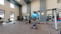 This well-appointed fitness center offers a comprehensive workout environment designed to accommodate a wide range of exercise disciplines. The space is defined by its soaring double-height ceilings accented with teal-painted beams, creating an open and energizing atmosphere throughout. Large black-framed floor-to-ceiling windows and elevated clerestory windows flood the interior with natural light while providing views of the surrounding exterior.

The main floor is finished with durable striped carpeting and is thoughtfully divided into distinct functional zones. A dedicated cardio area lines one end of the space, equipped with treadmills and elliptical machines. A strength and machine training zone features cable systems, weight benches, barbells, and a selection of selectorized equipment, complemented by full-length mirrors that enhance the sense of space. A functional training area includes a heavy punching bag, a rope ladder wall mount, plyo boxes, medicine balls, stability balls, a tire sled, and a dumbbell rack.

Wall-mounted sconce lighting adds warmth alongside the recessed ceiling fixtures, while a large flat-screen display serves the strength training area. Black-framed glass partition doors separate areas while maintaining visual continuity. The overall design balances industrial-modern aesthetics with practical, high-capacity fitness amenities.