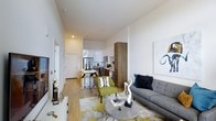This single-floor residence showcases a thoughtfully designed open-concept layout that integrates the living, dining, and kitchen areas into one cohesive space. The interior is characterized by crisp white walls and smooth ceilings with recessed lighting, creating a bright and airy ambiance throughout. Light-toned hardwood flooring runs continuously across the unit, contributing to a sense of warmth and continuity.

The living area is generously proportioned, anchored by a tufted gray sectional sofa and complemented by a vibrant green accent chair, a striped round coffee table, and a patterned area rug featuring yellow and gray tones. A large piece of wall art adds a contemporary artistic touch to the space.

The kitchen features a clean, modern design with white cabinetry, rich wood-toned tall storage units, and stainless steel appliances including a refrigerator and microwave. A peninsula with bar seating provides a casual dining option, illuminated by pendant lighting above. The overall layout ensures excellent sightlines between the kitchen and living areas, making the space feel open, connected, and well-suited for both everyday living and entertaining.