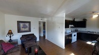 This single-floor residence offers a practical and comfortable open-concept layout that connects the living area and kitchen in one cohesive space. The living room features rich hardwood-style flooring that runs throughout, complemented by clean white walls that keep the space feeling bright and airy. A cozy seating area anchors the room, complete with a dark upholstered sofa and ottoman arranged around a sleek glass coffee table, with framed wall art adding a decorative touch. A half-wall partition subtly divides the living area from the kitchen, maintaining an open feel while defining each zone. The kitchen is equipped with dark cabinetry that provides a bold contrast against the white appliances, including an electric range with overhead ventilation. Ample counter space and additional storage make it a functional cooking area. A hallway visible from the living room leads to the bedroom, which offers a glimpse of additional living space beyond. Ceiling vents ensure climate control throughout, and natural light filters into the space, enhancing the warm, inviting atmosphere created by the hardwood flooring and neutral color palette.